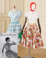 Two vintage-style dress mannequins displayed indoors against a paneled wall. The left mannequin wears a white dress with blue and black floral dots; the right mannequin wears a white blouse with a pink floral skirt and a red head covering.