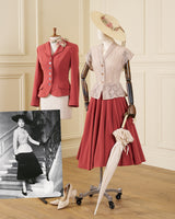 A vintage-style fashion display in an indoor room with cream paneled walls and wood flooring, featuring two mannequins dressed in coordinated outfits. One mannequin wears a coral-red blazer over a white top with a cream scarf, and the other wears a cream lace vest with a rust-red skirt, a cream hat, and holds a cream umbrella.