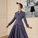 Woman in a vintage-style purple suit with a black hat, standing in a room with a framed black and white photo of another woman in a similar outfit.