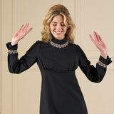 Woman wearing a black dress with decorative cuffs and neckline against a beige wall.