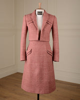 A pink and white checkered two-piece suit set consisting of a cropped blazer jacket and a matching knee-length skirt, displayed on a mannequin in a room with cream paneled walls and a wooden floor.
