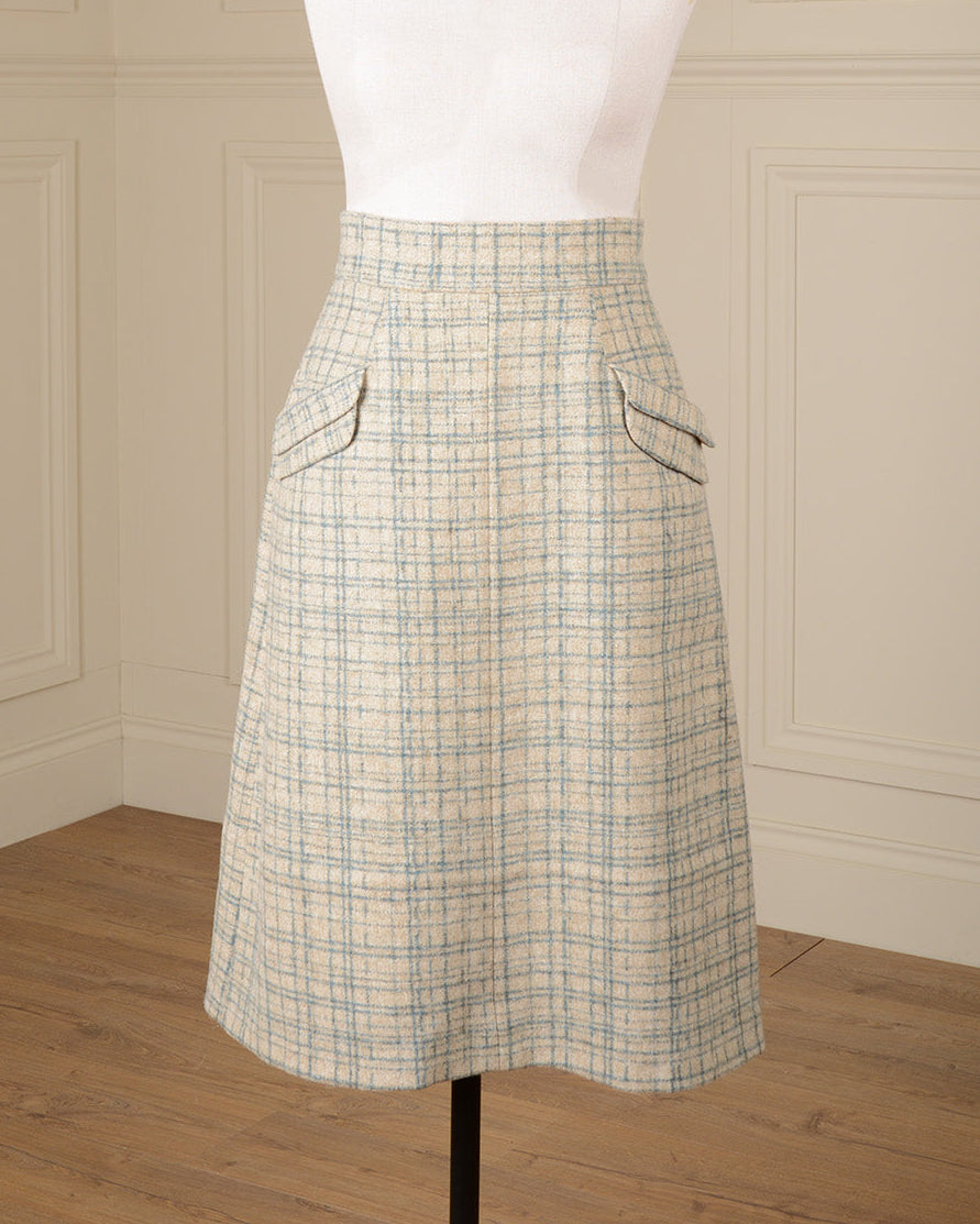 A high-waisted A-line skirt in a beige and light blue plaid tweed fabric, displayed on a white mannequin torso. The skirt features two angled front patch pockets and is shown against a background of cream paneled walls and a light wood floor.