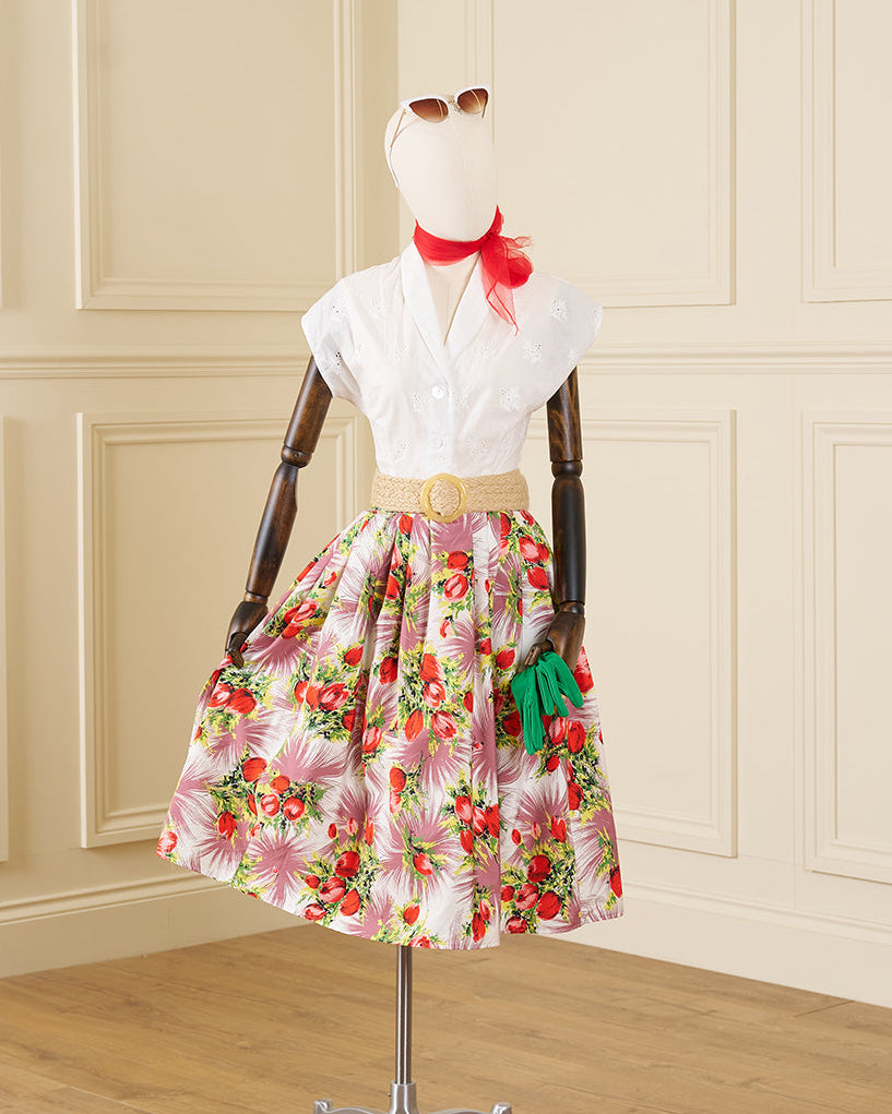 A vintage-style floral skirt dress displayed on a mannequin in an indoor room with cream paneled walls and a wood floor. The dress features a white short-sleeve blouse, a tan belt, and a full skirt with a pink, red, and green floral print. 