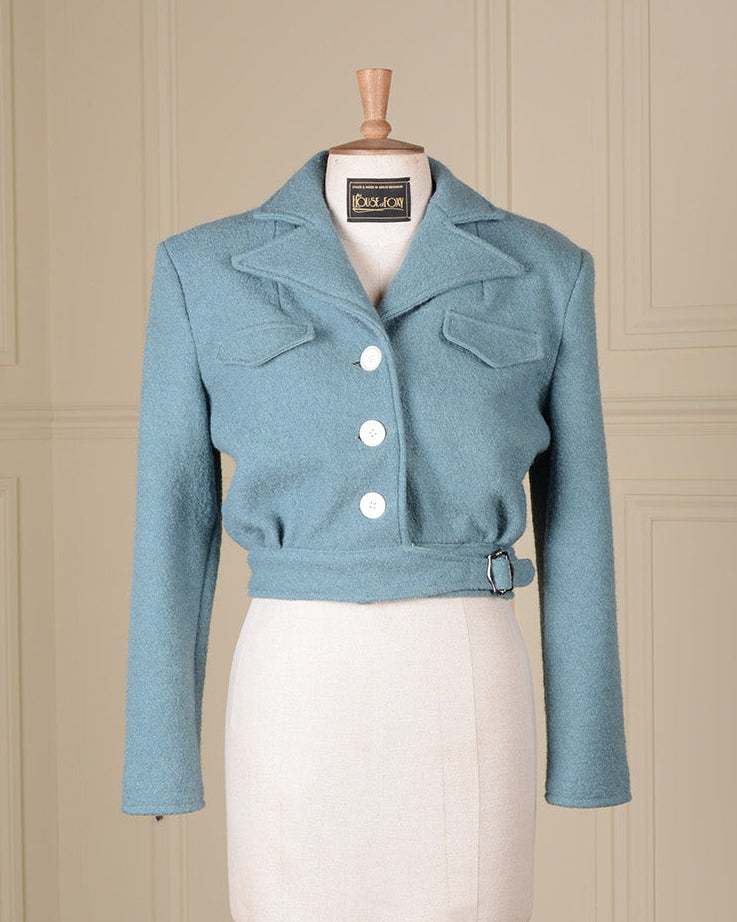 A light blue cropped jacket displayed on a cream-colored dress form mannequin, set against a beige paneled wall background.