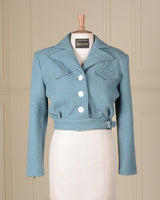 A light blue cropped jacket displayed on a cream-colored dress form mannequin, set against a beige paneled wall background.