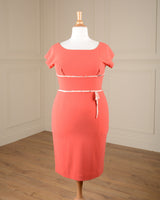 A coral pink short-sleeve knee-length dress displayed on a white mannequin in an indoor room with cream paneled walls and a wood floor.