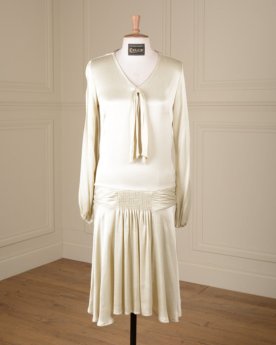 A cream-colored long-sleeve dress displayed on a mannequin in a room with cream paneled walls and a wooden floor. The dress features a V-neckline with a tie detail at the chest, a gathered waist, and a pleated skirt.