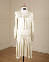 A cream-colored long-sleeve dress displayed on a mannequin in a room with cream paneled walls and a wooden floor. The dress features a V-neckline with a tie detail at the chest, a gathered waist, and a pleated skirt.