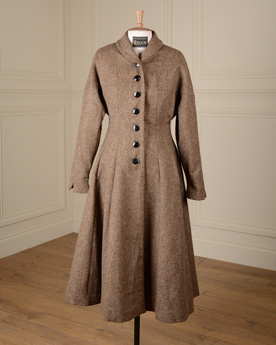 A brown tweed long coat with a high shawl collar and dark buttons, displayed on a dress form against a cream paneled wall and wood floor.