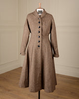 A brown tweed long coat with a high shawl collar and dark buttons, displayed on a dress form against a cream paneled wall and wood floor.