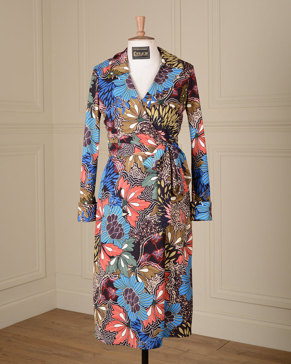 A long-sleeve wrap dress with a vibrant multicolor floral and abstract botanical print, displayed on a mannequin in front of a cream-colored paneled wall.