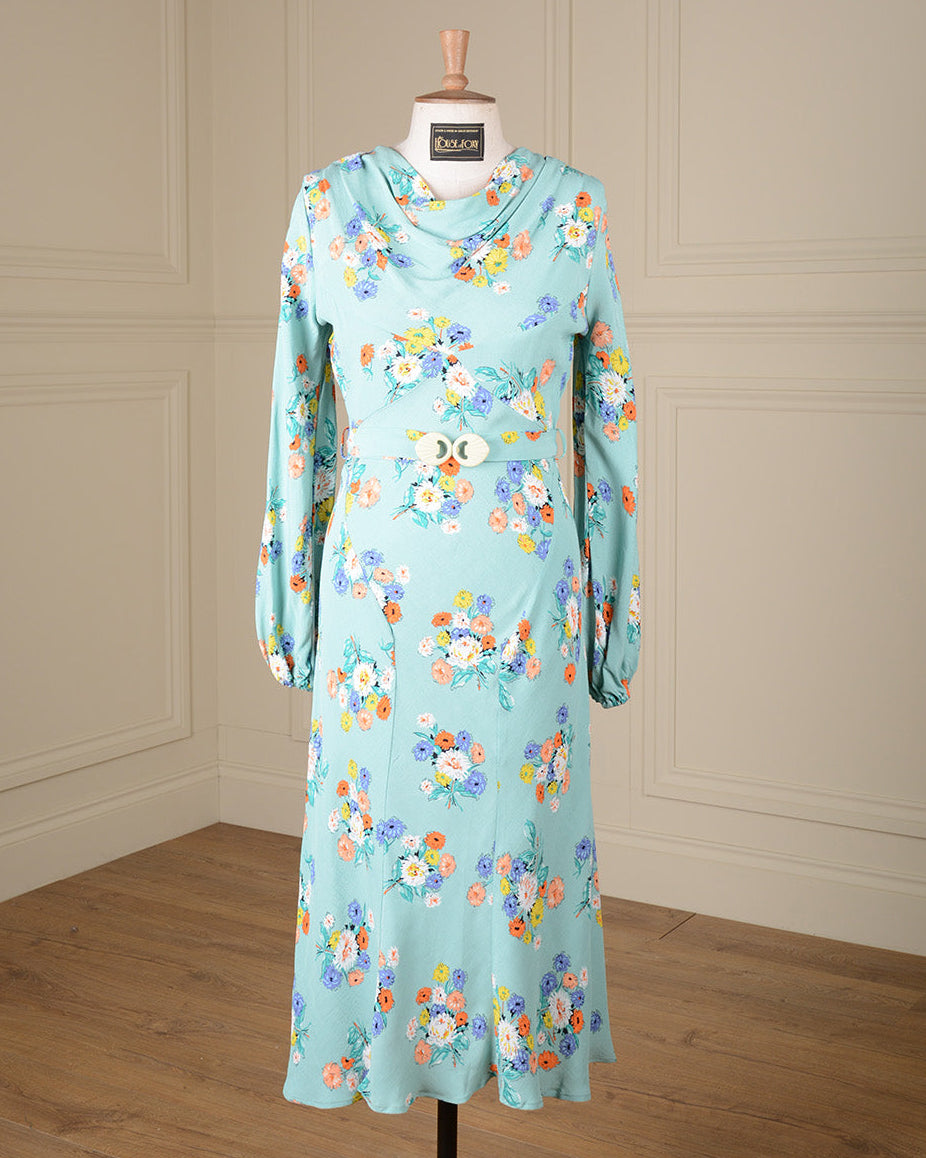 A light blue long-sleeve maxi dress with a colorful floral print, displayed on a mannequin in a room with cream paneled walls and a wooden floor.
