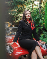 A woman in a black vintage-style suit with a red scarf sits on the hood of a red classic car, with green foliage in the background.