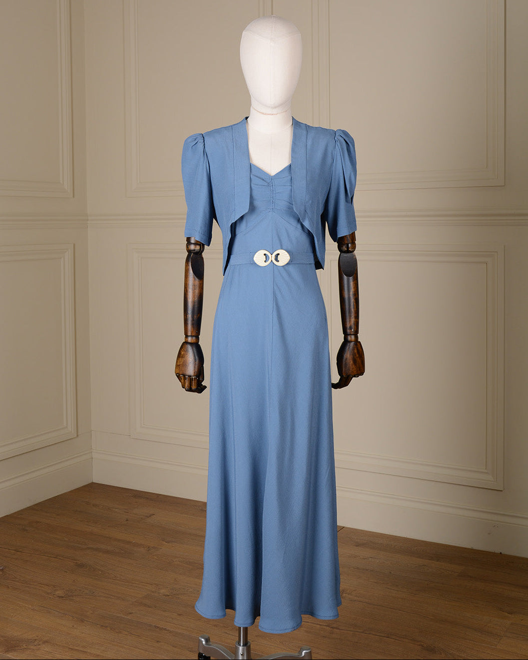 A light blue vintage-style dress displayed on a mannequin with wooden arms, set against a cream paneled wall and wood floor