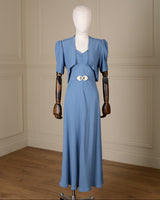 A light blue vintage-style dress displayed on a mannequin with wooden arms, set against a cream paneled wall and wood floor