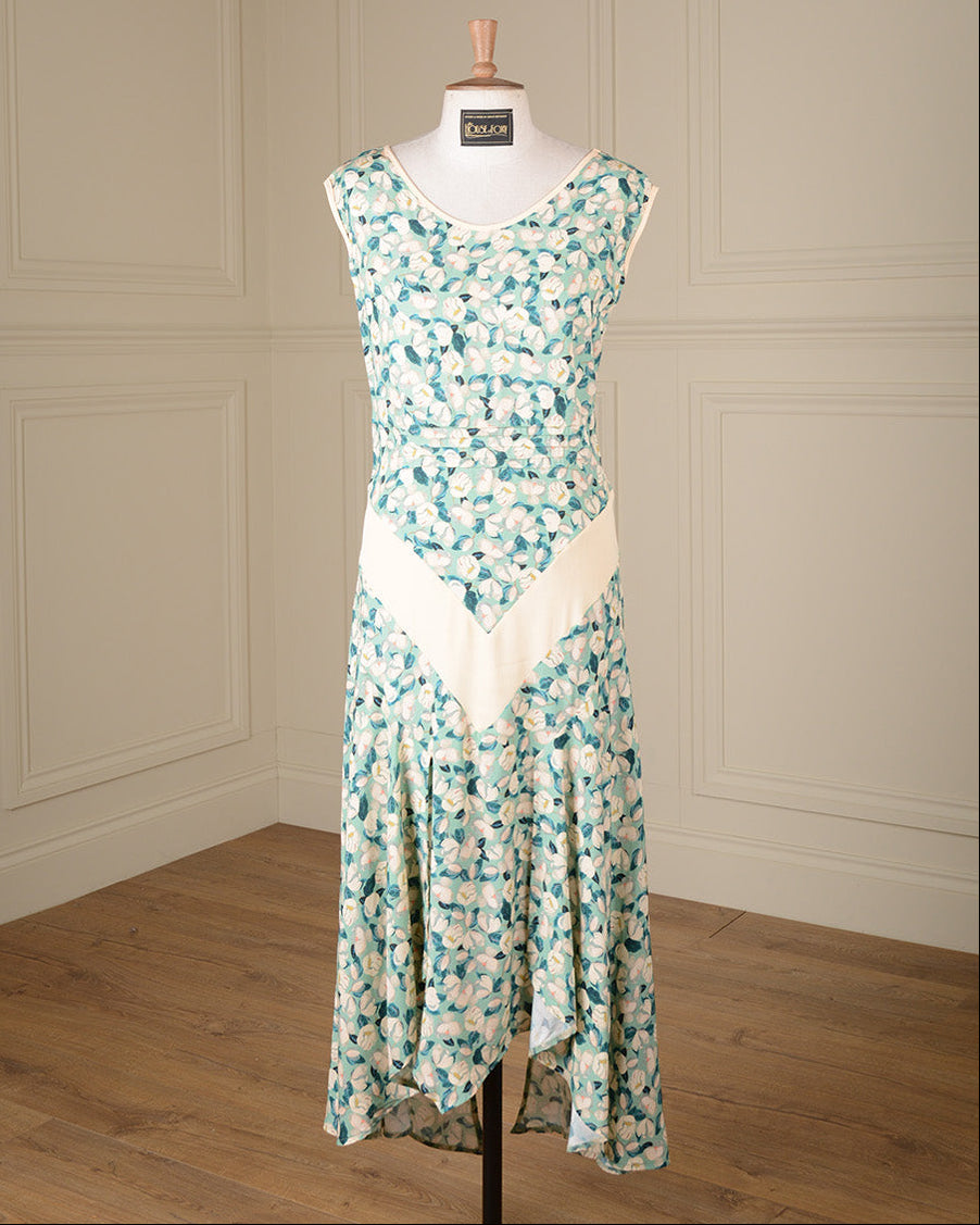 A sleeveless V-neck dress displayed on a mannequin in an indoor room with cream paneled walls and a wood floor. The dress has a teal and white floral print with a contrasting solid cream V-shaped panel at the waist