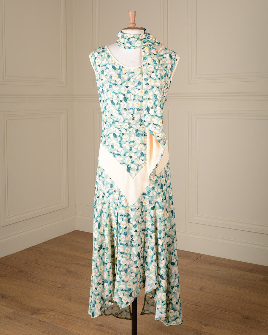 A sleeveless high-neck maxi dress with a green and white floral print and a cream-colored wrap-style front panel, displayed on a mannequin in a room with cream paneled walls and a wooden floor.