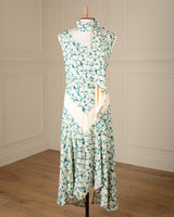 A sleeveless high-neck maxi dress with a green and white floral print and a cream-colored wrap-style front panel, displayed on a mannequin in a room with cream paneled walls and a wooden floor.