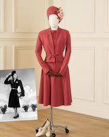 A vintage-style terracotta red two-piece skirt suit displayed on a mannequin, featuring a long-sleeve jacket with a tie waist and matching pleated skirt. The outfit is styled with a matching floral fascinator hat, a small brooch, and a pair of beige heels on a rolling stand, set against a cream paneled wall background with a black-and-white inset photo of a model wearing the outfit.