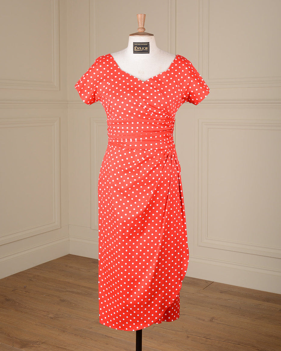 A red short-sleeve dress with white polka dots displayed on a white mannequin torso in a room with cream paneled walls and a wood floor.