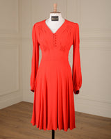 A bright red long-sleeve midi dress displayed on a mannequin in an indoor room with cream paneled walls and a wood floor. The dress has a V-neckline with decorative buttons, gathered detailing at the shoulders and bust, and a pleated skirt