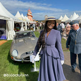 A woman wearing a lavender purple two-piece suit and a wide-brimmed straw hat stands next to a classic silver convertible car at an outdoor event. A man in a military uniform walks nearby, and white tents are visible in the background under a blue sky.