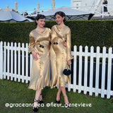 Two women posing outdoors in matching shiny gold midi dresses with long sleeves and black belt details, standing in front of a white picket fence and green hedges.