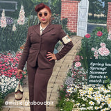 A woman stands in a garden-themed setting wearing a brown tailored suit with a white collared shirt, round sunglasses, and holding a small handbag. A brick wall, white gate, and flower beds are visible in the background, along with the text 'Spring has Spring! Summer florals to match! garden.