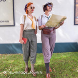 Two women dressed in vintage 1950s travel-style clothing walking outdoors on grass, one holding a red clutch and the other a large paper map. The outfits feature muted gray, lavender, beige, and yellow tones with brown accessories.