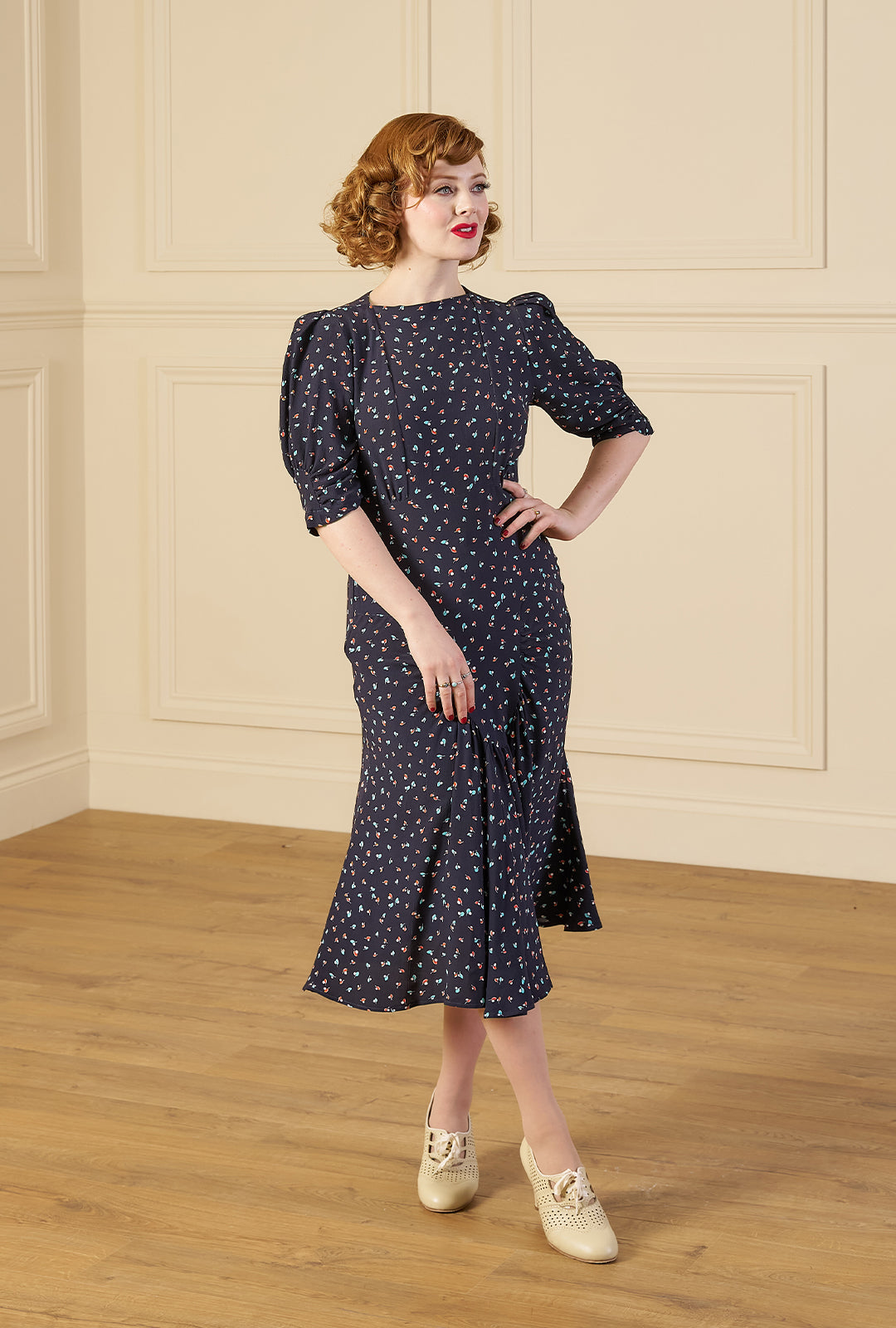Woman wearing a 1940s print tea midi dress in ocean pearl navy dark blue dress with white patterns in a room with wooden floor and paneled walls.
