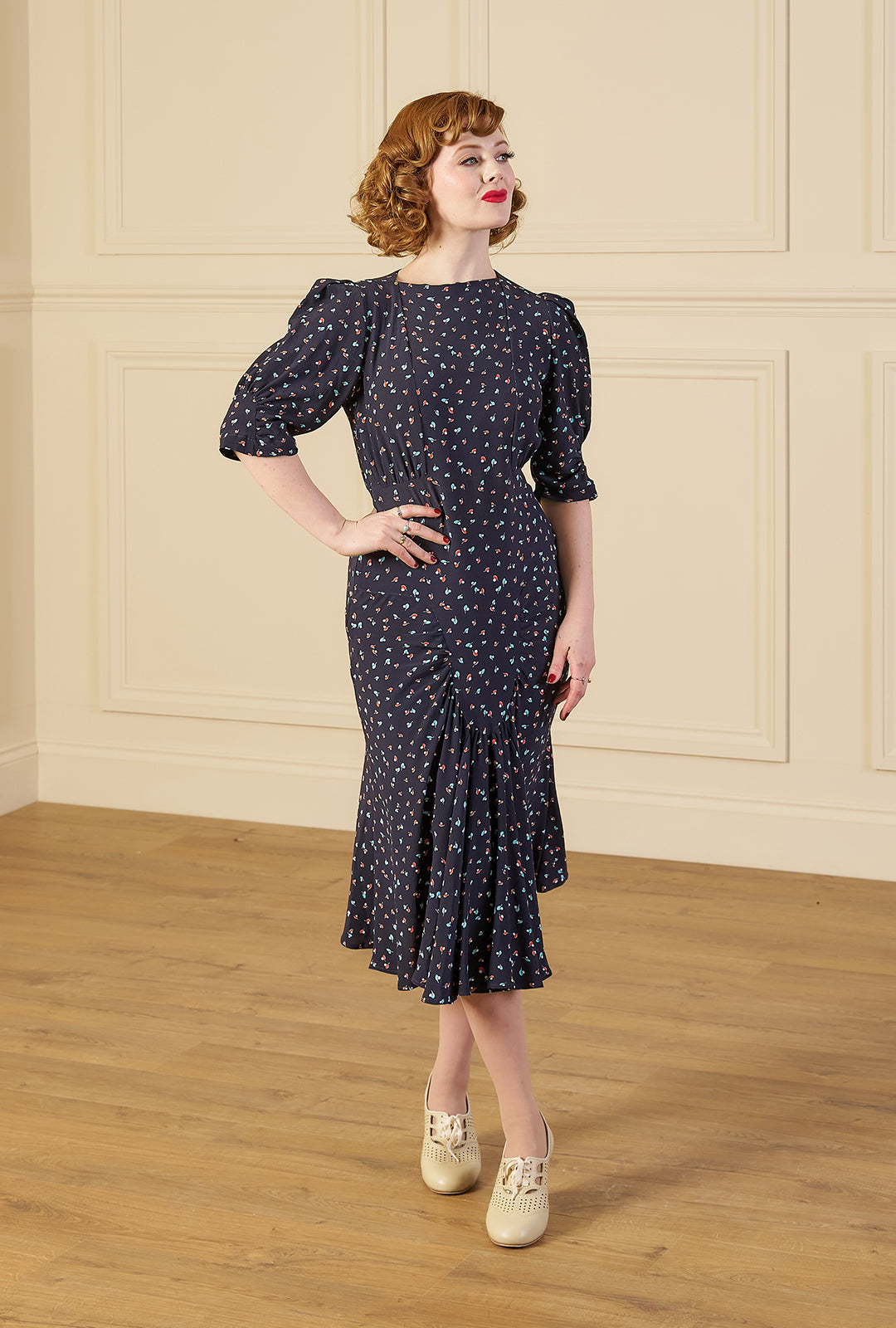 Woman wearing a 1940s print tea midi dress in ocean pearl navy dark blue dress with white patterns in a room with wooden floor and paneled walls.