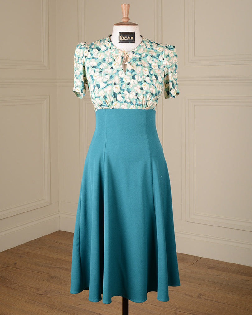 A vintage-style two-piece dress displayed on a mannequin in a room with cream paneled walls and a wood floor. The top has a white base with a green floral pattern and short puff sleeves, while the skirt is a solid teal color.