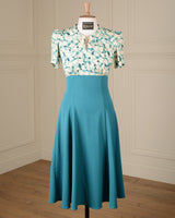 A vintage-style two-piece dress displayed on a mannequin in a room with cream paneled walls and a wood floor. The top has a white base with a green floral pattern and short puff sleeves, while the skirt is a solid teal color.
