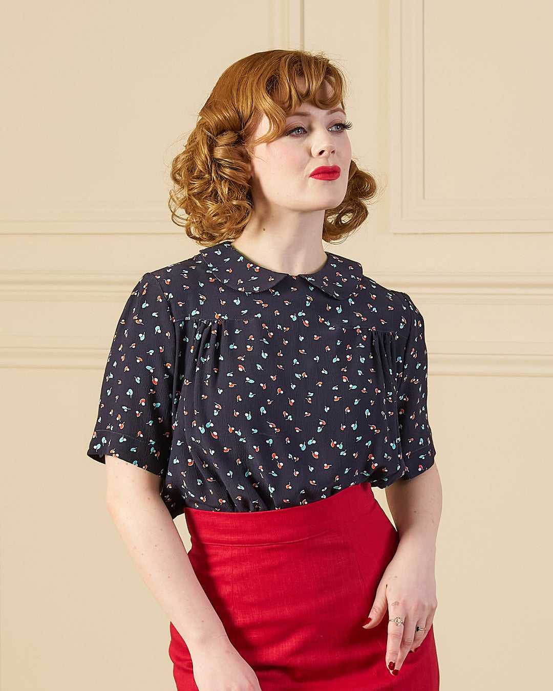 Woman wearing a black patterned top and red skirt against a beige wall.