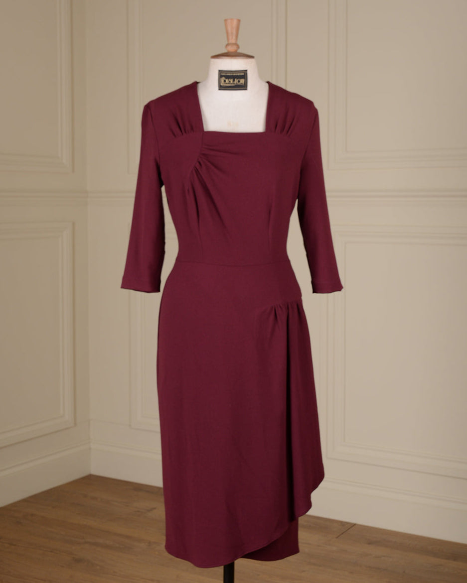 A knee-length burgundy dress with three-quarter sleeves and a square neckline, displayed on a white mannequin torso in an indoor room with cream paneled walls and a wood floor.