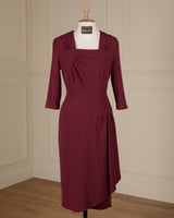 A knee-length burgundy dress with three-quarter sleeves and a square neckline, displayed on a white mannequin torso in an indoor room with cream paneled walls and a wood floor.