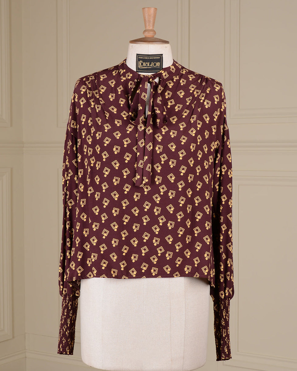 A long-sleeve blouse with a bow tie neckline, featuring an all-over repeating geometric pattern in beige on a deep burgundy base, displayed on a cream dress form