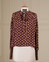 A long-sleeve blouse with a bow tie neckline, featuring an all-over repeating geometric pattern in beige on a deep burgundy base, displayed on a cream dress form