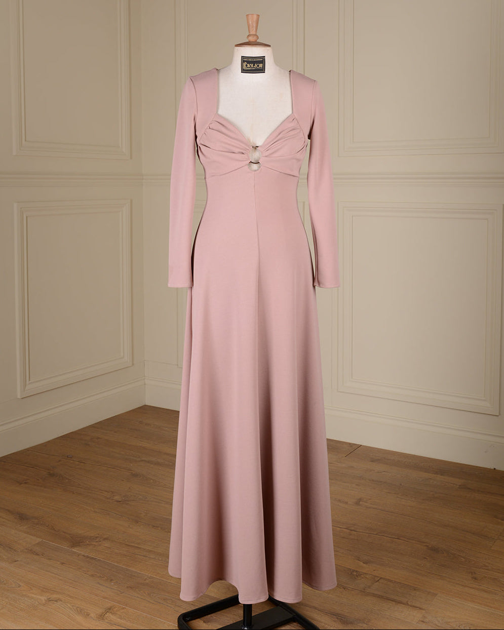A floor-length dusty pink gown displayed on a white mannequin in a room with cream paneled walls and a wood floor. The dress features long sleeves, a square neckline, a sweetheart-style bust with gathered draping and a central decorative button detail, and a fitted silhouette that flows into a full skirt.