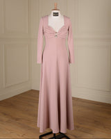 A floor-length dusty pink gown displayed on a white mannequin in a room with cream paneled walls and a wood floor. The dress features long sleeves, a square neckline, a sweetheart-style bust with gathered draping and a central decorative button detail, and a fitted silhouette that flows into a full skirt.