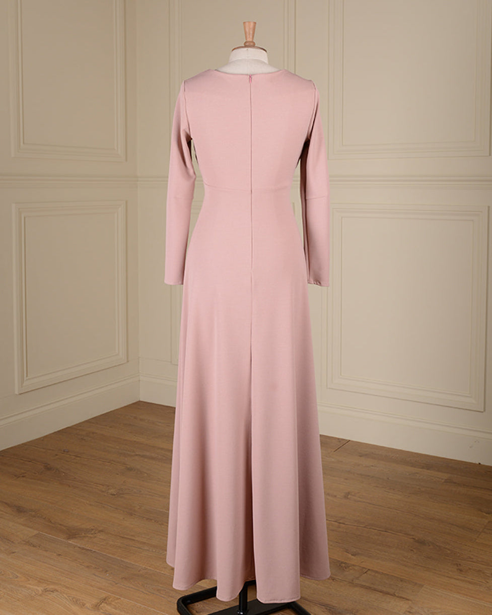 A long-sleeved dusty pink maxi dress displayed on a mannequin from the back, set against a cream paneled wall and light wood floor.