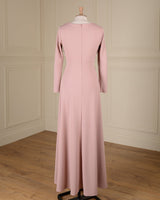 A long-sleeved dusty pink maxi dress displayed on a mannequin from the back, set against a cream paneled wall and light wood floor.